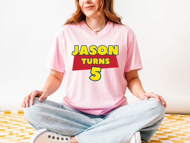 May include: A light pink short-sleeved t-shirt featuring a graphic design with the yellow text JASON, the red text TURNS, and the large yellow number 5. The shirt is paired with light blue denim jeans and white sneakers.