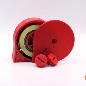 Könnte beinhalten: Ein roter Plastik-Klebebandspender mit einer Rolle gelben und grünen Klebebands. Der Spender hat einen runden Deckel und zwei kleine rote Plastikteile.