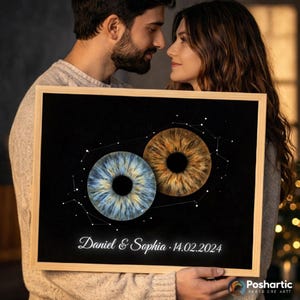 May include: A framed print with two iris illustrations, one blue and one brown, on a black background. The print displays the names "Daniel & Sophia" and the date "14.02.2024" in script, with a constellation design. The frame is made of light wood.