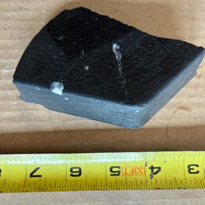 May include: A dark gray, roughly rectangular object with a textured surface. The object is approximately 4 inches long and 3 inches wide. There are white spots and a white line on the surface. The object is on a brown surface.