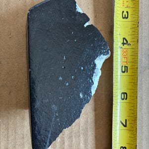 May include: A black, irregularly shaped piece of material with a rough, textured surface. The piece is approximately 4 inches long. A yellow measuring tape is positioned next to the object.