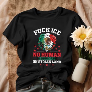 Pu&ograve; includere: T-shirt nera con la scritta "FUCK ICE" sopra una grafica della bandiera messicana, un'aquila e una rosa rossa. Sotto, si legge "NO HUMAN IS ILLEGAL ON STOLEN LAND". La maglietta &egrave; su una superficie beige con fiori.