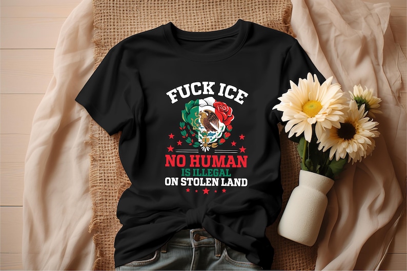 Pu&ograve; includere: T-shirt nera con la scritta "FUCK ICE" sopra una grafica della bandiera messicana, un'aquila e una rosa rossa. Sotto, si legge "NO HUMAN IS ILLEGAL ON STOLEN LAND". La maglietta &egrave; su una superficie beige con fiori.