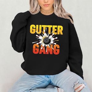 May include: Black sweatshirt with the text "GUTTER GANG" in yellow and orange. A bowling ball is surrounded by white and red bowling pins. The sweatshirt is a casual apparel item.