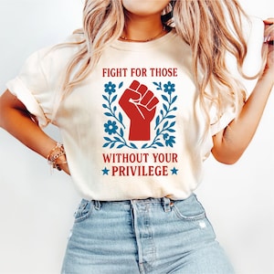 May include: Cream-colored t-shirt featuring a red fist graphic surrounded by blue floral elements. The text "FIGHT FOR THOSE WITHOUT YOUR PRIVILEGE" is printed above and below the fist. The shirt is made of a soft material.