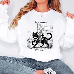 May include: White sweatshirt featuring a black and white graphic of a knight riding a black cat. The text reads "Defend Your Local" and "public library". The design is a vintage style illustration.