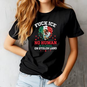 Pu&ograve; includere: T-shirt nera con la scritta "FUCK ICE" sopra "NO HUMAN IS ILLEGAL ON STOLEN LAND". Il design include lo stemma messicano, una rosa rossa e dettagli verdi e bianchi. La maglietta &egrave; indossata da una persona.