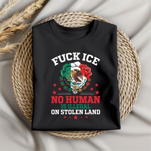 Pu&ograve; includere: T-shirt nera con la scritta "FUCK ICE" sopra una grafica della bandiera messicana e una rosa rossa. Sotto, il testo recita "NO HUMAN IS ILLEGAL ON STOLEN LAND". La maglietta &egrave; piegata su una superficie tessuta.
