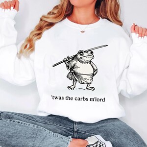 Pode incluir: Camisola branca com um desenho de uma rã guerreira segurando uma espada. A rã está vestida com uma túnica e tem uma expressão severa. O texto "'twas the carbs m'lord" está impresso abaixo.