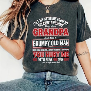 Może przedstawiać: Ciemnoszara koszulka z napisem "I GET MY ATTITUDE FROM MY FREAKIN' AWESOME GRANDPA" w kolorze białym i czerwonym. Koszulka zawiera również napisy "GRUMPY OLD MAN" i "YOU HURT ME AND THEY'LL NEVER FIND YOUR BODY".