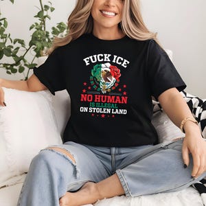 Pu&ograve; includere: T-shirt nera con la scritta "FUCK ICE" sopra una grafica della bandiera messicana, una rosa e un'aquila. Sotto, la maglietta recita "NO HUMAN IS ILLEGAL ON STOLEN LAND". La maglietta &egrave; indossata da una persona.