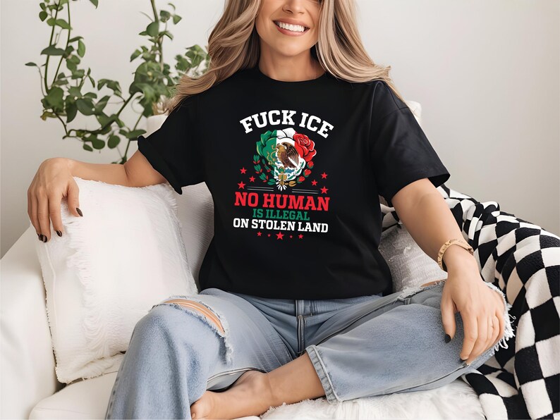 Pu&ograve; includere: T-shirt nera con la scritta "FUCK ICE" sopra una grafica della bandiera messicana, una rosa e un'aquila. Sotto, la maglietta recita "NO HUMAN IS ILLEGAL ON STOLEN LAND". La maglietta &egrave; indossata da una persona.