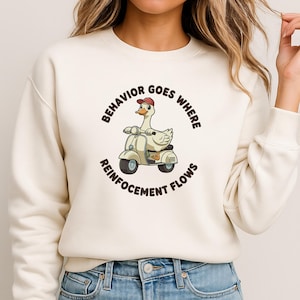 May include: A cream-coloured sweatshirt with a cartoon goose on a scooter, wearing a red cap. The text "BEHAVIOR GOES WHERE REINFORCEMENT FLOWS" is printed around the goose. The sweatshirt is a casual, comfortable style.