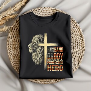 May include: Black t-shirt featuring a lion's profile and a cross design. The text reads "HUSBAND DADDY PROTECTOR HERO" in a patriotic color scheme. The shirt is folded and placed on a woven basket.