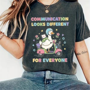 May include: Dark grey t-shirt with the text "COMMUNICATION LOOKS DIFFERENT FOR EVERYONE". The shirt features a cartoon goose riding a pink scooter, wearing a helmet and carrying a laptop.