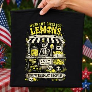 May include: A black fabric item featuring a graphic print of a skeleton wearing a cap at a lemonade stand. The text reads: WHEN LIFE GIVES YOU LEMONS, THROW THEM AT PEOPLE. The design includes yellow lemons, a jar, a crate, and skulls in yellow and white.