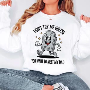 May include: White sweatshirt with the text "DON'T TRY ME UNLESS YOU WANT TO MEET MY DAD" and a cartoon tombstone character. The tombstone is gray with a smiling face and is running. Gold stars are around the tombstone.