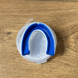 May include: A blue and white mouth guard in a clear plastic case. The mouth guard is U-shaped and designed to protect teeth. Instructions are printed on the case.