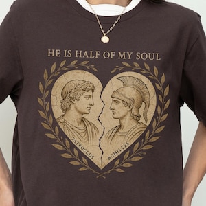 Könnte beinhalten: Dunkelbraunes T-Shirt mit weißem Kragen. Das Design zeigt zwei klassische Figuren in Herzform mit dem Text "HE IS HALF OF MY SOUL". Die Figuren sind mit Patroclus und Achilles beschriftet.