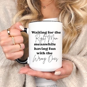 May include: White ceramic mug with a black handle and rim. The mug features the text "Waiting for the Right Man meanwhile having fun with the Wrong Ones" in black script and block letters. The mug is held by a person.