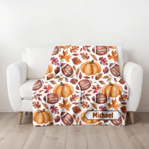 May include: A white blanket with a repeating pattern of watercolor pumpkins, footballs, and autumn leaves in shades of orange, brown, and red. The blanket has the name "Michael" in a decorative frame.