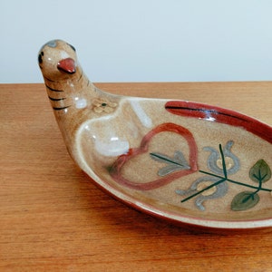 May include: A ceramic bird-shaped dish with a tan base colour. The dish features a red beak, black stripes on the neck, and decorative floral and heart-shaped designs in red, green, and white. The dish is approximately 30 cm long.