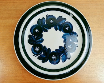 Könnte beinhalten: Ein runder, weißer Keramikteller mit dunkelgrünem Rand und floralem Design. Der Teller zeigt einen Kranz aus blauen Blumen mit dunkelblauen Akzenten. Der Teller steht auf einer hellbraunen Holzoberfläche.