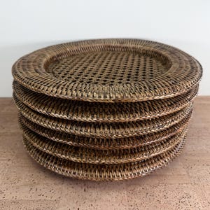 May include: A stack of six round, woven rattan chargers. The chargers are a natural brown colour with a woven design and a perforated centre. The chargers are stacked on top of each other.