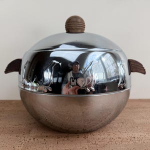 Puede incluir: Una cubitera plateada con tapa y asa de madera. La cubitera tiene forma redonda con una superficie brillante y reflectante. Presenta diseños de pingüinos alrededor de la base y asas de madera en los laterales. La tapa tiene un pomo de madera.