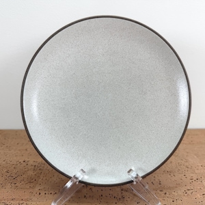 Puede incluir: Un plato redondo de cerámica con una superficie de color gris claro moteado y acabado liso. El plato presenta un borde fino y distintivo de color marrón oscuro que rodea todo el contorno exterior, contrastando con el centro más claro.