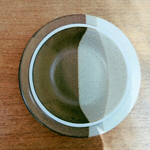 May include: Overhead view of a ceramic bowl with a segmented design. The bowl features a dark brown, light brown, and speckled white color scheme, separated by a white band. The bowl is set against a wooden surface.