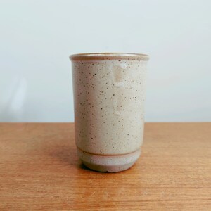 May include: A speckled ceramic tumbler with a light beige glaze and a slightly flared rim. The cylindrical cup has a rounded base and is set against a wooden surface.