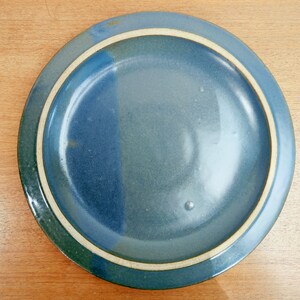 May include: A round ceramic plate with a blue and green color scheme. The plate has a white rim and a glossy finish. The plate is on a wooden surface.