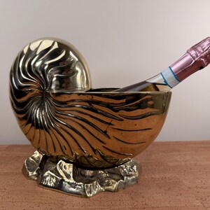 May include: A gold-colored, nautilus-shaped wine chiller with a bottle of champagne. The chiller has a detailed, spiral design and sits on a decorative base. The champagne bottle has a pink foil top.