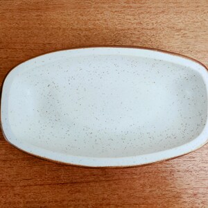 May include: An oval-shaped, off-white ceramic serving dish with a speckled surface. The rim is a dark brown color. The dish is empty and sits on a wooden surface.