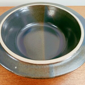 May include: A ceramic bowl with a wide, flat rim and a deep interior. The bowl is primarily a muted blue-green color, with a lighter, off-white band around the inner rim. The exterior rim has a darker, mottled appearance. The bowl is likely for serving.