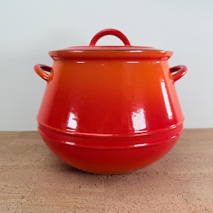 May include: A vibrant, red-orange enameled cast iron pot with a lid. The pot has two side handles and a loop handle on the lid. The pot is sitting on a wooden surface.