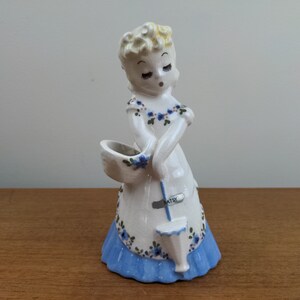 May include: A vintage ceramic figurine of a woman in a white dress with blue floral accents and a blue skirt. The figurine holds a basket and a small container. The word "Betsy" is on the apron.