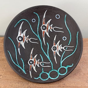 May include: Round, dark brown ceramic plate with a stylized fish design. The fish are white with orange and black details, set against a background of teal seaweed and bubbles. A decorative piece.