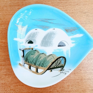 May include: A vintage, teardrop-shaped ashtray with a light blue background and a winter scene. The scene depicts two igloos, a sled, and the text "Out Am". The ashtray is made of ceramic or glass.