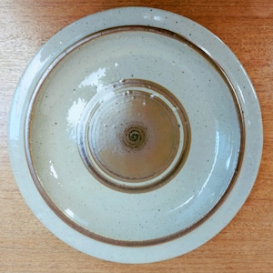 May include: Overhead view of a ceramic plate with a light beige glaze and speckled texture. The plate features concentric brown rings and a central, recessed area. The rim has a raised edge with a brown border. The plate is on a wooden surface.
