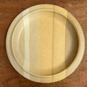 May include: A round ceramic plate with a raised rim. The plate features vertical stripes of varying shades of yellow and beige. The speckled surface adds texture to the dish, suitable for serving food.