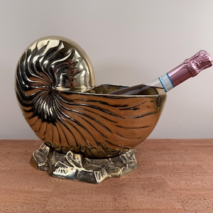 May include: A gold-colored, nautilus shell-shaped wine chiller with a bottle of sparkling wine inside. The chiller sits on a decorative base. The shell design features detailed ridges and curves.
