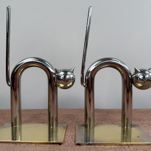 May include: A pair of chrome cat-shaped bookends. Each bookend features a curved body, a cat head with round eyes, and a tail. The base is a square of gold-colored metal. The bookends are designed to stand upright.