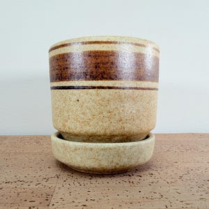 May include: A ceramic planter with a matching saucer. The planter is cylindrical with a speckled beige finish and two brown bands. The saucer is a matching speckled beige. This decorative item is suitable for indoor plants.