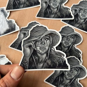 Puede incluir: Pegatinas en blanco y negro con una ilustración detallada de una persona con un sombrero de ala ancha, con arte facial y cabello largo. Las pegatinas tienen el texto "@old_evvel".