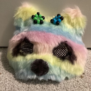 May include: A colorful, furry panda mask in pastel rainbow hues. The mask features black mesh eye coverings, a dark brown nose, and two beaded accents on top. The fur is a blend of yellow, pink, blue, and green.