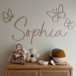 May include: A wooden cabinet with a woven front holds a variety of items. Above, the name "Sophia" is spelled out in cursive, accompanied by wooden butterfly cutouts. A small basket, flower-shaped pillow, and decorative ball are on top.