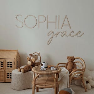 Può includere: Un cartello in legno marrone chiaro con il nome "Sophia grace" scritto su di esso. Il cartello è appeso a un muro bianco. In primo piano c'è una piccola casetta di bambole in legno, un giocattolo di peluche e un tavolo e sedie in rattan.