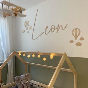 Puede incluir: Una cama de madera con forma de casa con un juguete de peluche de elefante gris y un parachoques de cuerda trenzada gris. La pared detrás de la cama está pintada de verde y tiene un letrero de nombre de madera que dice "Leon" con globos aerostáticos y nubes.